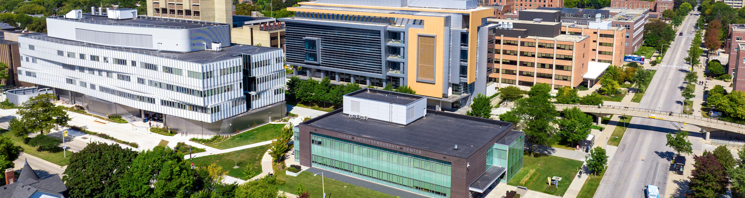 Aerial view of the UW-Milwaukee campus on a sunny day, featuring the Lubar Entrepreneurship Center in the foreground and several modern academic buildings surrounded by green trees and walkways.