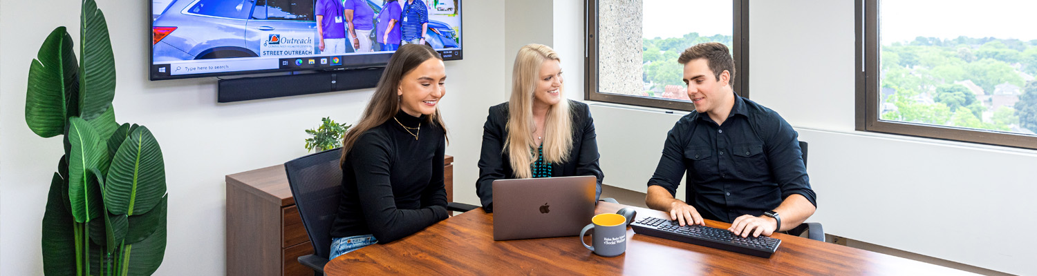 Three colleagues smile and collaborate around a wooden conference table with a laptop and keyboard, in a bright modern office with large windows and a wall-mounted screen displaying a street outreach program in the background.