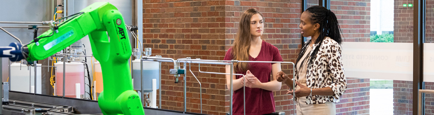 Student and Professor work together in an engineering lab, examining equipment near a green industrial robotic arm against an exposed brick wall.