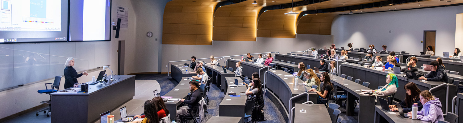 A professor lectures to a large group of students in a modern university lecture hall with curved tiered seating, warm wood-paneled walls, and a projected presentation screen at the front of the room.