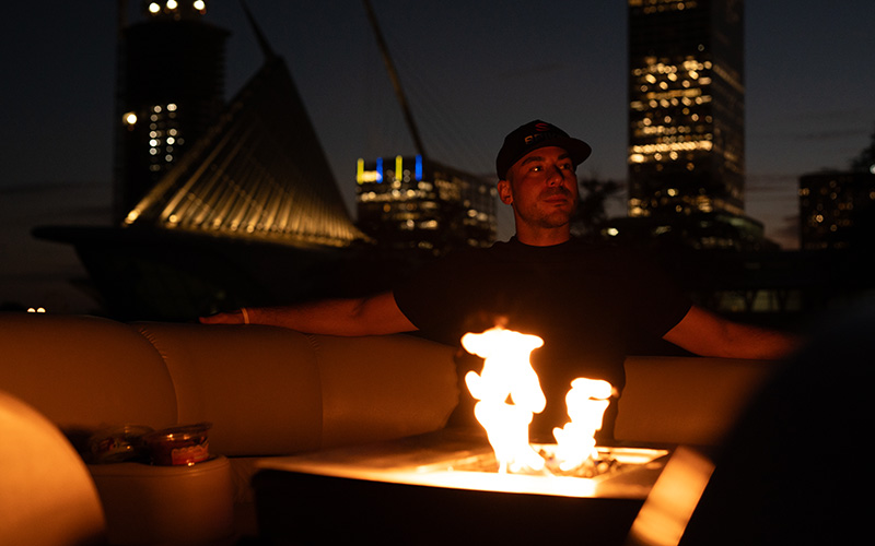 Ben Caya sits in front of a fire with the Milwaukee skyline behind him.
