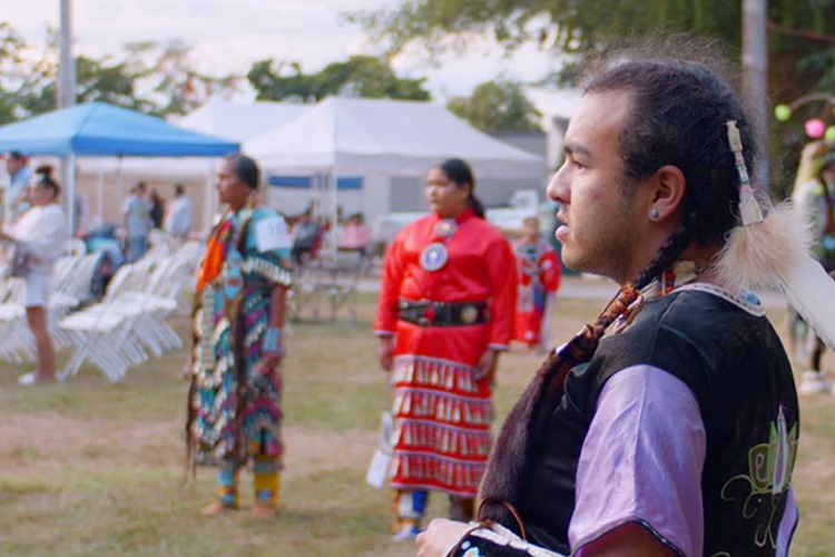 A still image from the documentary film Powwow people. Three people stand on grass watching something off camera.