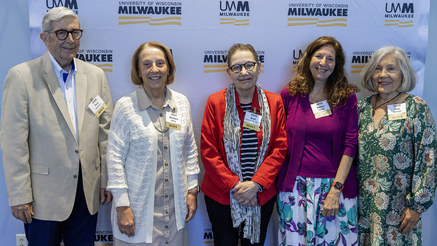 Five volunteers wearing "HOST" name badges stand in front of a University of Wisconsin–Milwaukee branded backdrop, smiling for a group photo. The group includes one man in a light-colored blazer and four women dressed in a mix of neutral and brightly colored outfits, including a red jacket, magenta cardigan, and green floral dress.