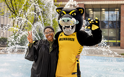 A graduate and Pounce pose for a photo in front of the UWM fountain