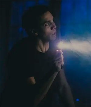 Travis A. Knight holds a flashlight in the darkness onstage with a concerned look on his face.