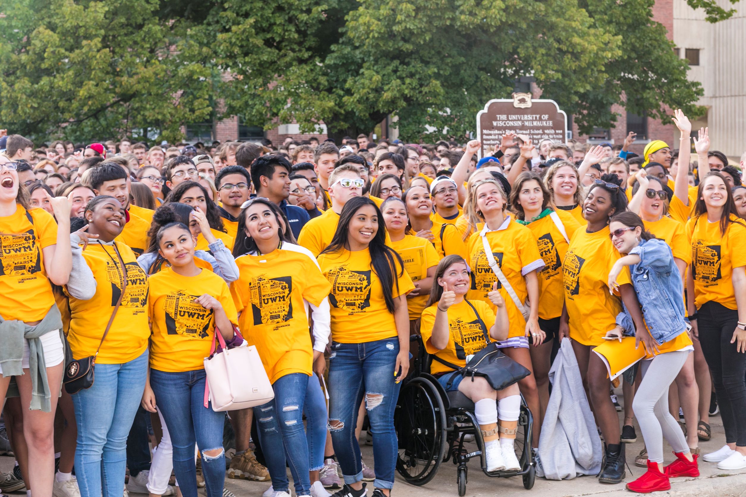 Large diverse crowd of UWM students smiling in front of Mitchell Hall during Welcome Week. Foreground female student in wheelchair giving thumbs up.
