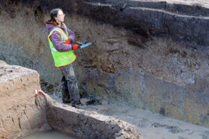 Archaeologist in a safety vest recording observations while standing in a deep excavation trench with visible soil layers