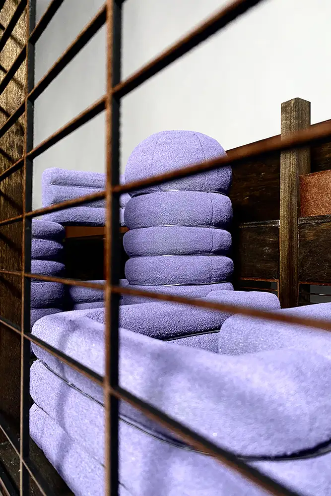 Close-up view through a wooden slatted screen of stacked, lavender-colored cushioned seating or folded textiles arranged on a wooden model.