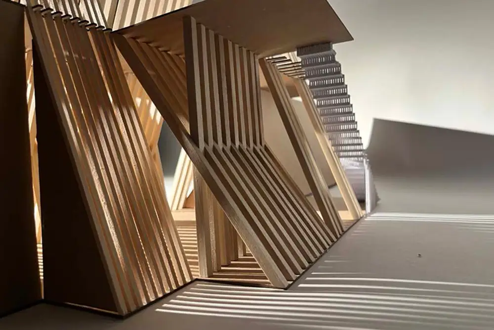 Close-up of a wooden architectural model with angled slatted panels and structural frames, casting striped shadows across the floor under warm light.
