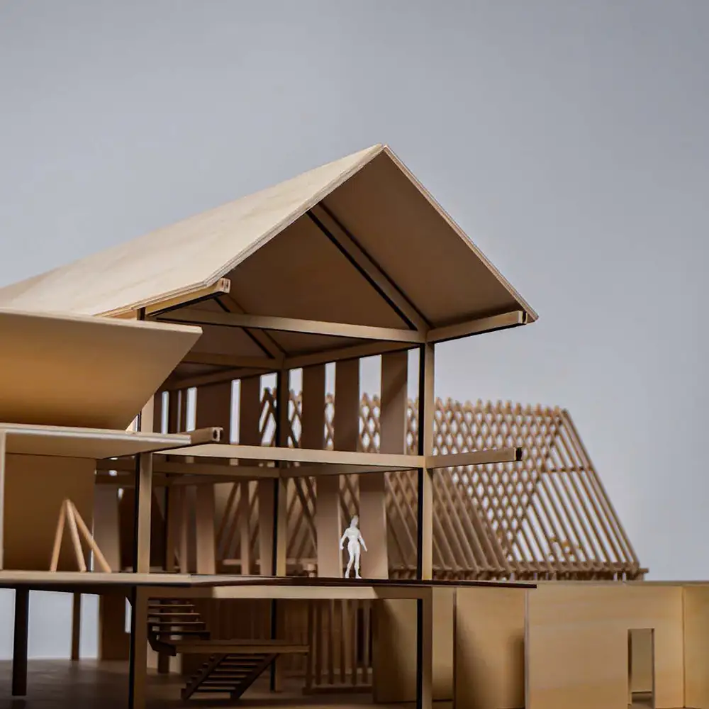 Cutaway architectural model of a wood-framed building with a pitched roof, lattice façade, interior stairs, and a small figure for scale.