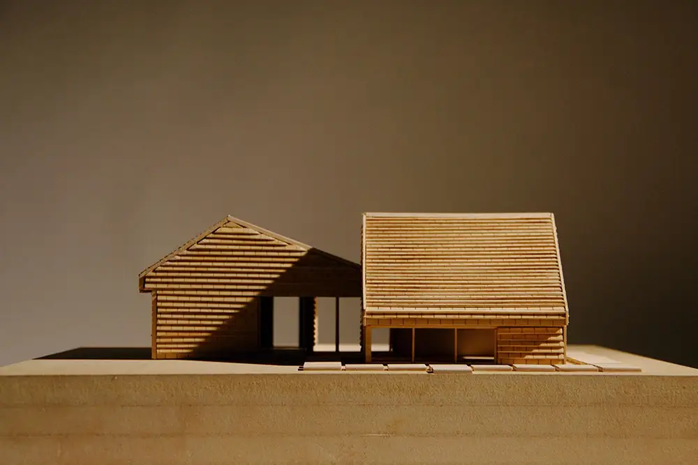 Front view of a wooden architectural model with two pitched‑roof volumes, horizontal slat cladding, and a small connecting entry, lit against a neutral backdrop.