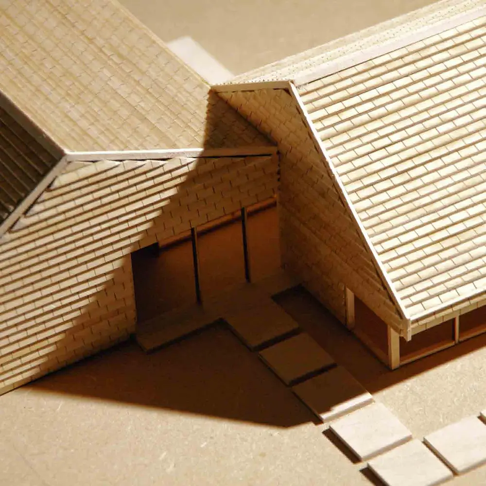 Close-up of a wooden architectural model showing intersecting pitched roofs, layered shingle details, recessed windows, and a shaded entry courtyard.