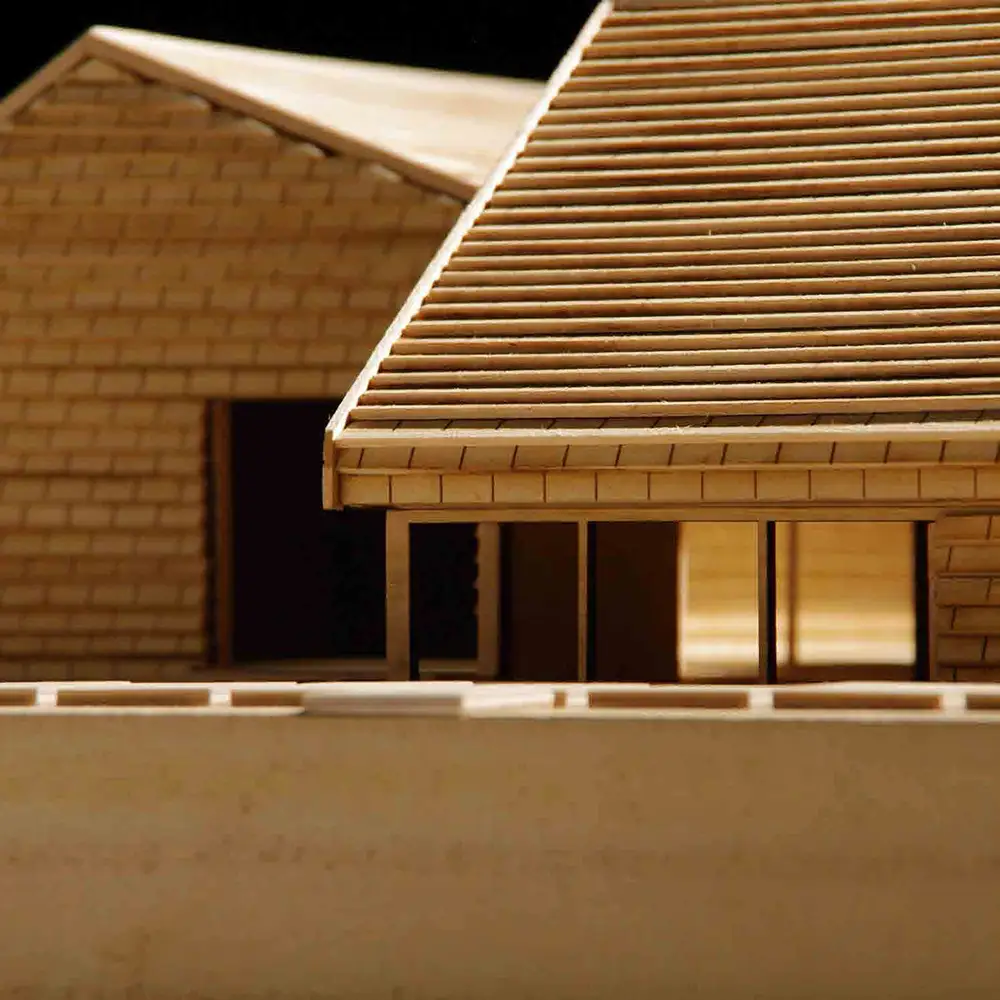 Close-up of a wooden architectural model highlighting horizontal roof slats, eaves, and window openings, with layered textures and warm studio lighting.