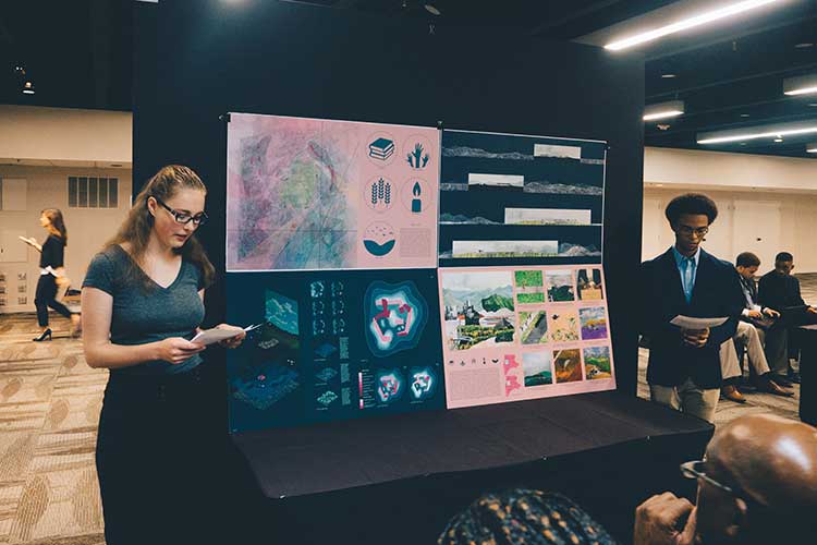 Architecture students in formal attire are seen at a table with posters of their work.