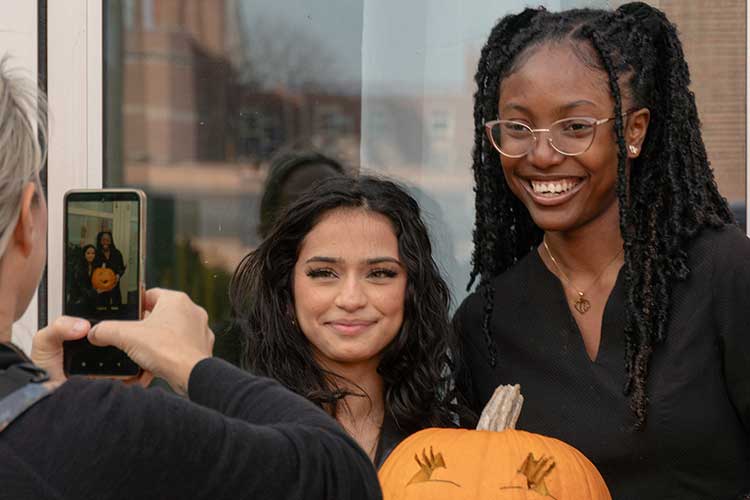 Two students get their picture taken while holding a pumpkin they carved during a social event.