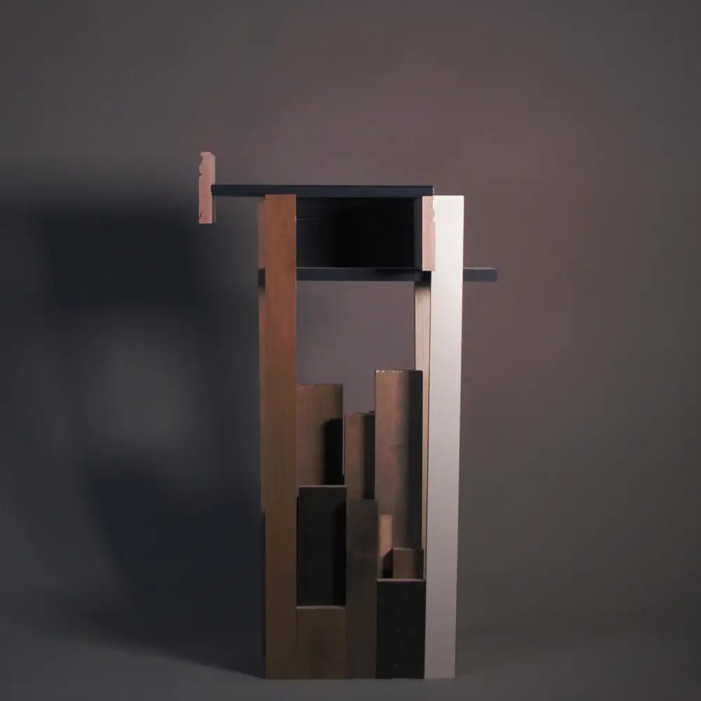 Abstract architectural model with vertical wooden and dark elements forming a narrow tower-like composition, topped by a horizontal black slab against a gray backdrop.