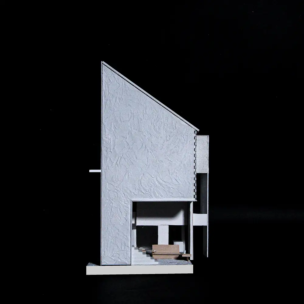 Section model of a compact house with a steeply angled roof and textured exterior, cut open to reveal interior stair, platform, and seating against a black background.