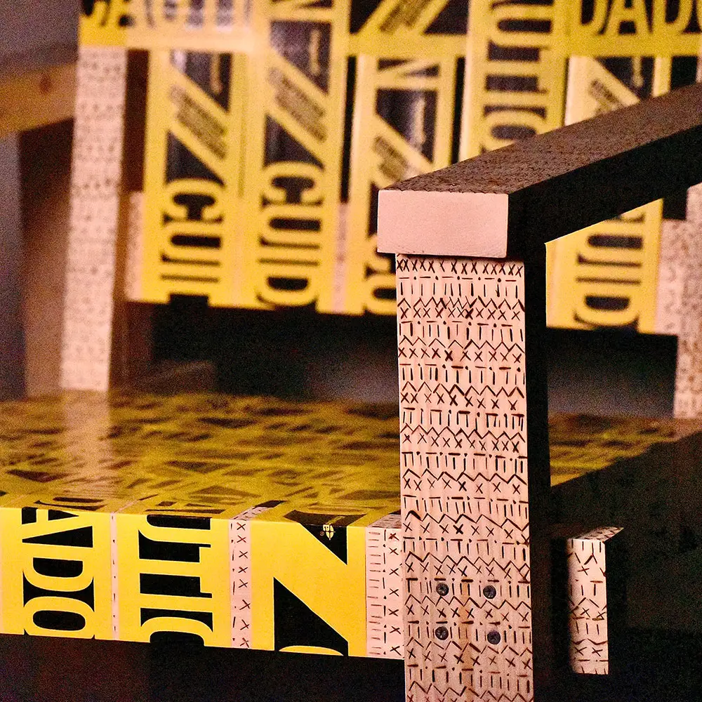 Close-up of a handcrafted wooden chair, showing layered plywood legs and surfaces wrapped in yellow caution tape with bold black lettering.