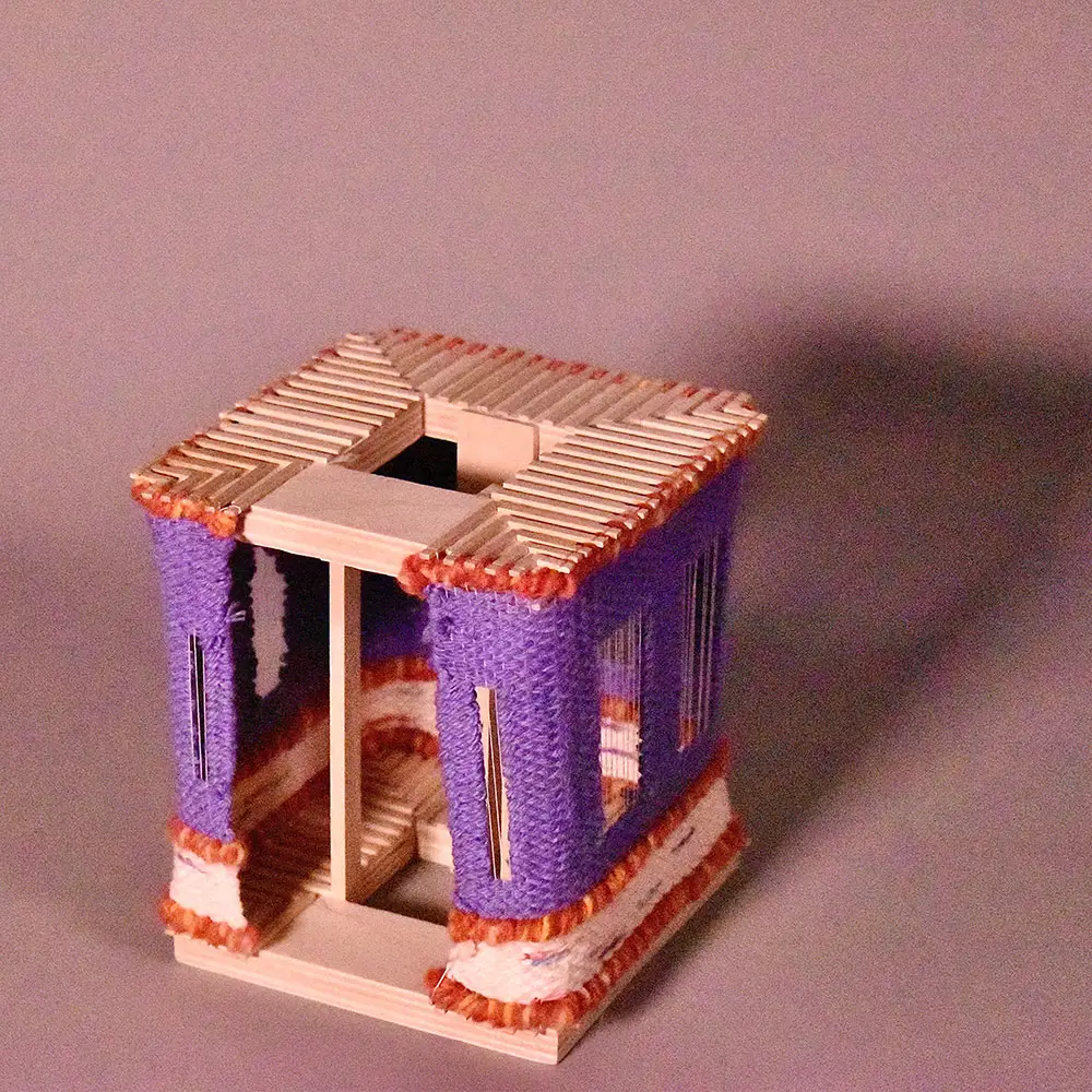 Small handcrafted cube sculpture made of wood and woven yarn, with purple panels, orange accents, and a hollow center, photographed against a plain backdrop.