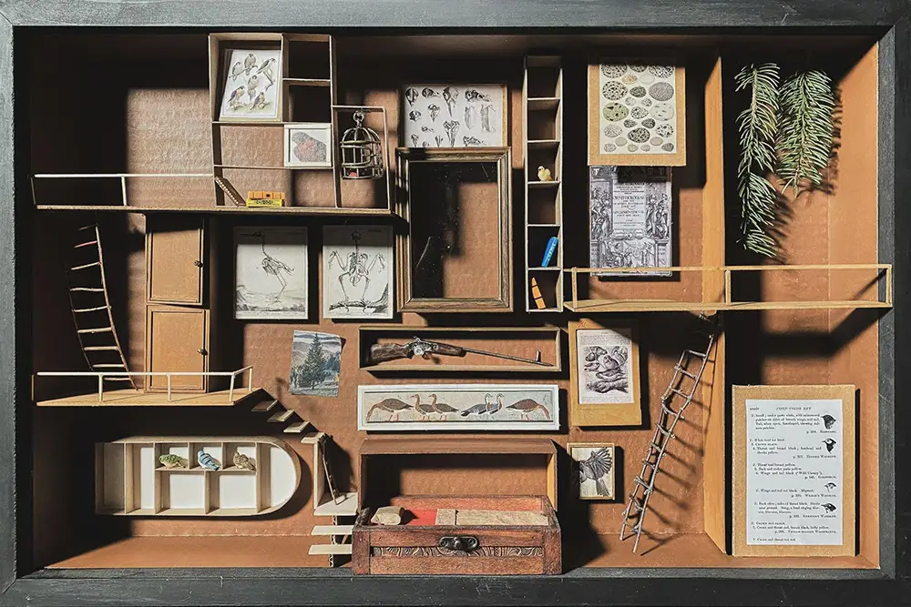 Overhead view of a boxed architectural assemblage with layered wooden compartments holding framed drawings, books, ladders, and small sculptural objects.