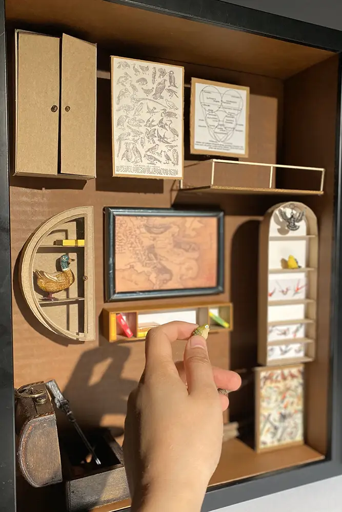 Hand interacting with a boxed wooden assemblage of miniature shelves, cabinets, framed illustrations, and small objects arranged like an interior display.