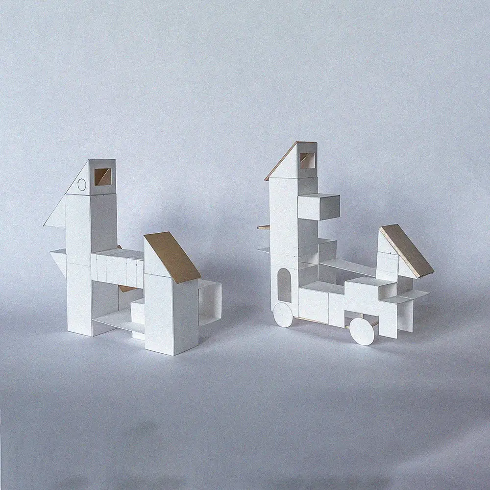 Two small white architectural models resembling abstract bird forms, built from stacked geometric blocks with pitched roofs, set on a neutral surface.