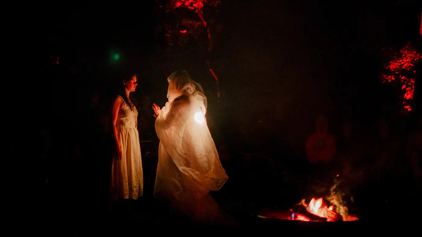 Two figures in flowing white garments stand near a fire in a dark outdoor setting with red lights.