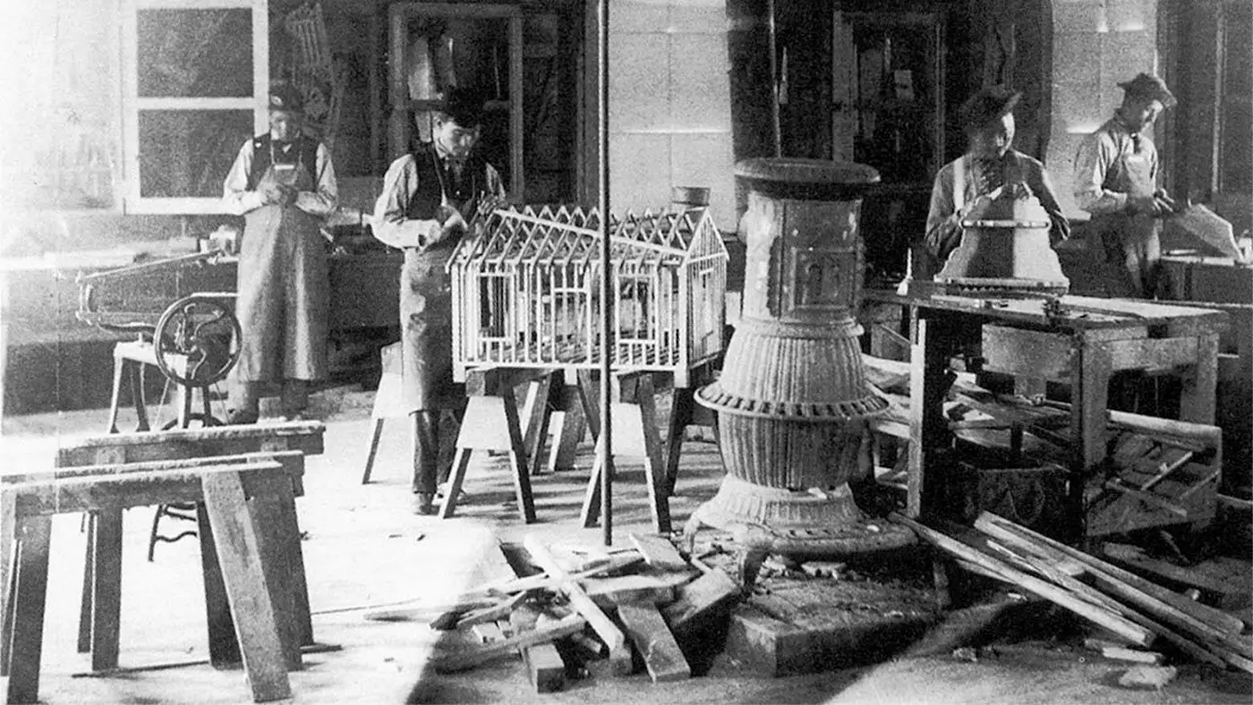 A black and white photo depicting a homebuilding course in the late 1800s. Several figures in aprons work with wood and other materials.