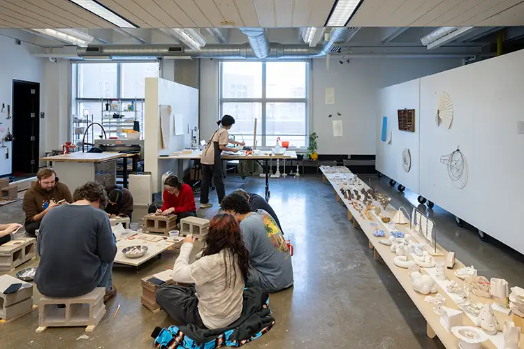 A group of students sit in a circle working with their hands on clay projects. Iris is in the background.