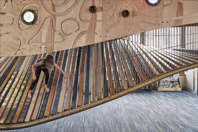 Child climbing a slanted ramp of wooden slats inside a designed play space, with curved railings, patterned wood panels, and soft indoor lighting. Child climbing a slanted ramp of wooden slats inside a designed play space, with curved railings, patterned wood panels, and soft indoor lighting.