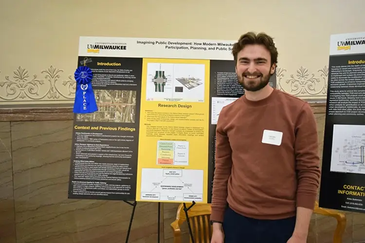 A young man with brown hair and a beard smiles while standing in front of a research poster, which has earned a blue ribbon.