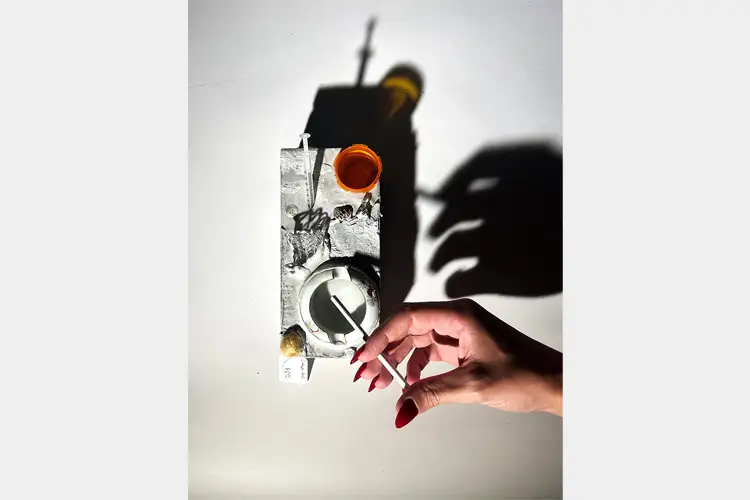 Hand holding a cigarette over a metal ashtray and objects, casting strong shadows on a white surface.