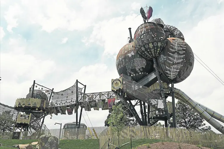 Large sculptural playground with giant circular towers, patterned surfaces, rope bridges, and a slide under a cloudy sky. Large sculptural playground with giant circular towers, patterned surfaces, rope bridges, and a slide under a cloudy sky.