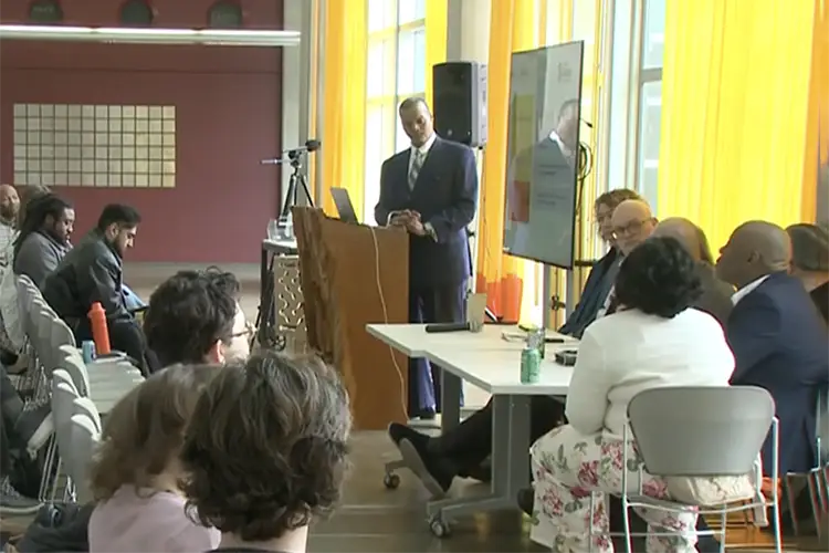 Dr. Kirk Harris stands at a podium addressing a panel of experts in the Marcus Commons.