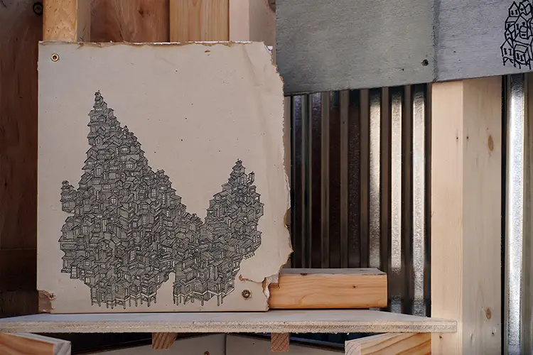 Wood panel with a drawing of clustered house-like forms, mounted within a rough wooden frame.
