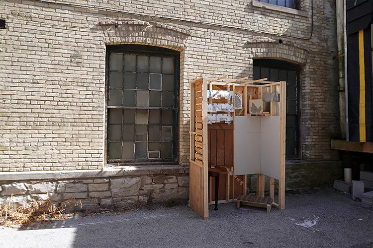 Small wooden structure installation placed in an alley against a brick wall with large windows.