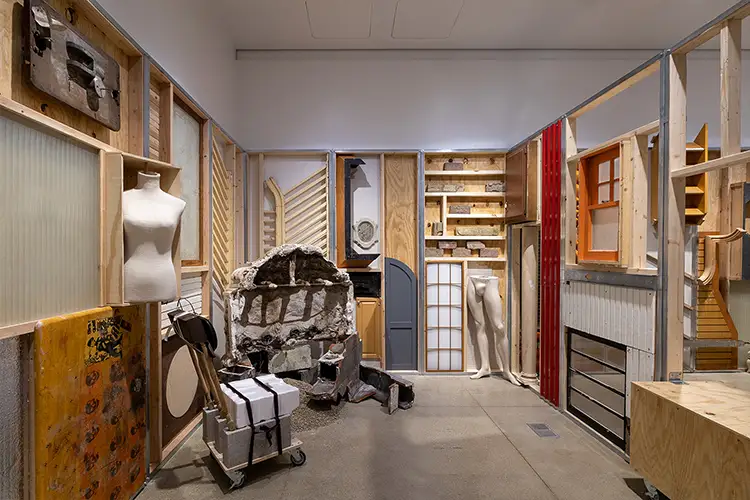 Interior installation of wood panels, sculptural fragments, and mannequins arranged like a workshop.