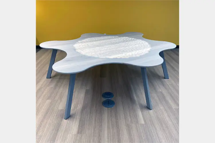 Low, irregularly shaped white table with blue legs, featuring a textured circular center, set on wood flooring against a yellow wall. Low, irregularly shaped white table with blue legs, featuring a textured circular center, set on wood flooring against a yellow wall.