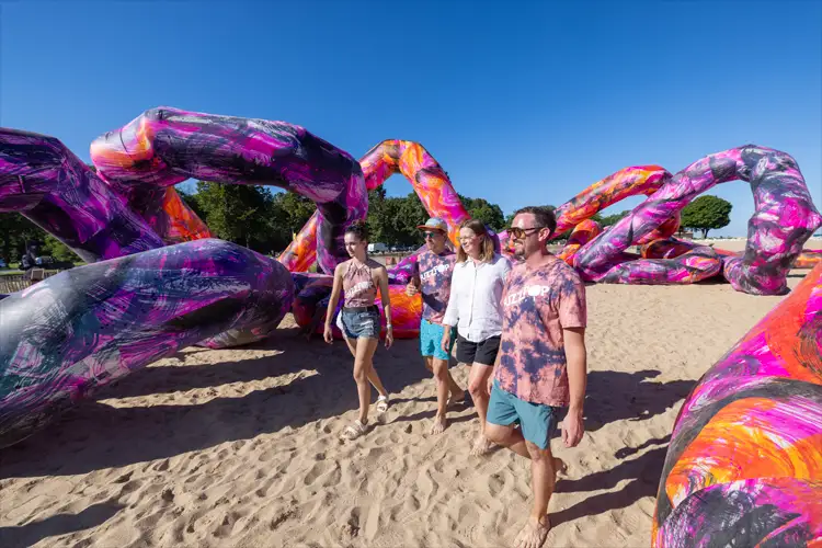 Four people walk barefoot on sand between large, colorful abstract inflatable sculptures with swirling pink, purple, and orange patterns under a blue sky. Four people walk barefoot on sand between large, colorful abstract inflatable sculptures with swirling pink, purple, and orange patterns under a blue sky.