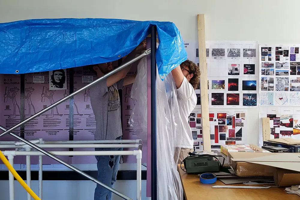Two people adjusting a blue tarp over a tall framed structure in a studio, with pipes, tools, and architectural posters on the wall behind.