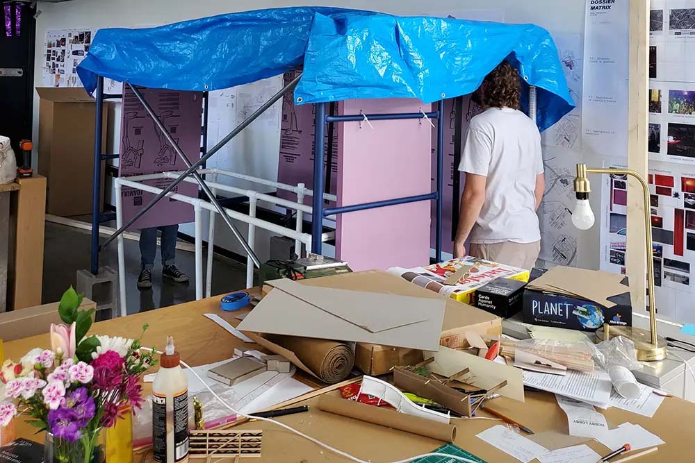 Studio scene with two people working under a blue tarp on a small scaffold structure; table in foreground cluttered with tools, cardboard, and materials.