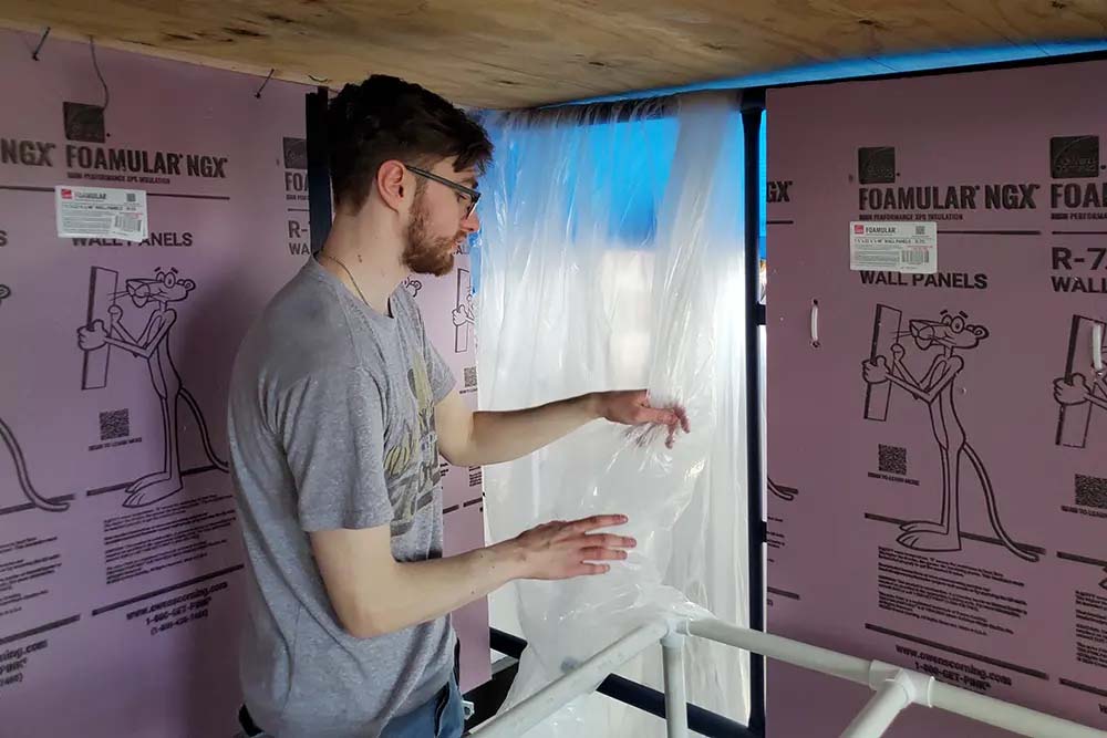 Interior architectural model view of a person holding a sheet of clear plastic inside a framed structure, surrounded by pink foam panels and pipes.
