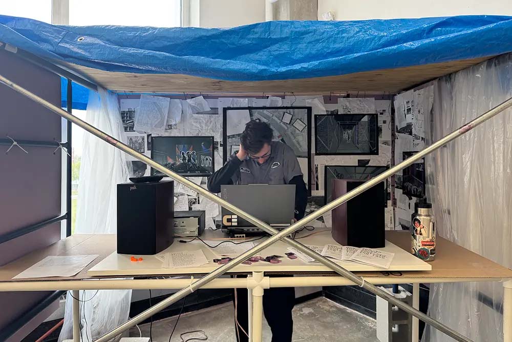 Person working at a desk under a blue tarp–covered frame, with speakers, papers, and pinned drawings on the walls behind.