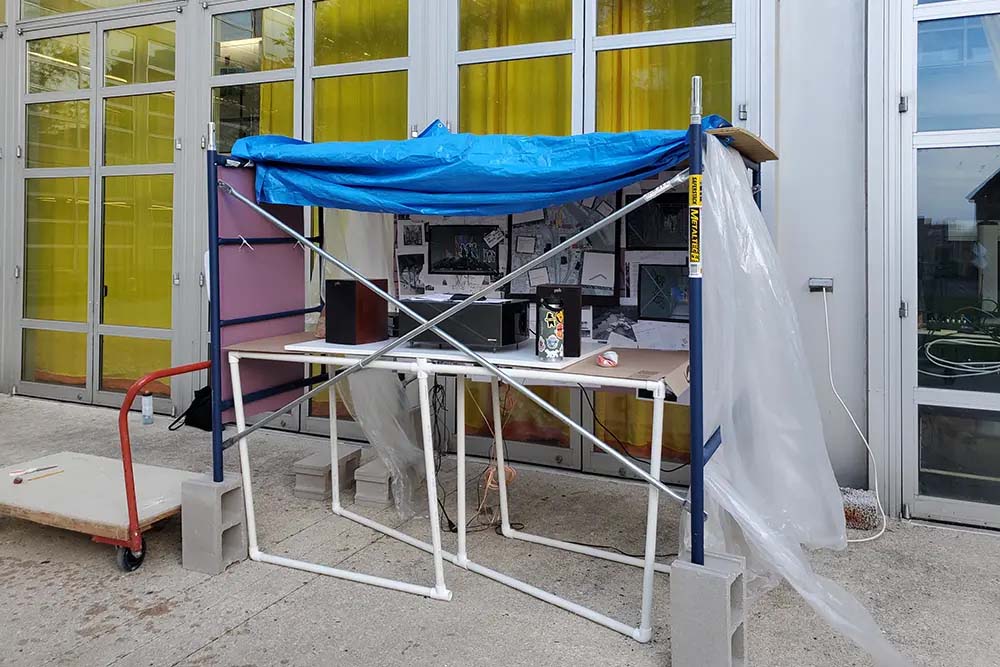 Outdoor view of a small scaffold-like structure with blue tarp roof, plastic sheeting, and desk setup in front of glass doors with yellow panels.