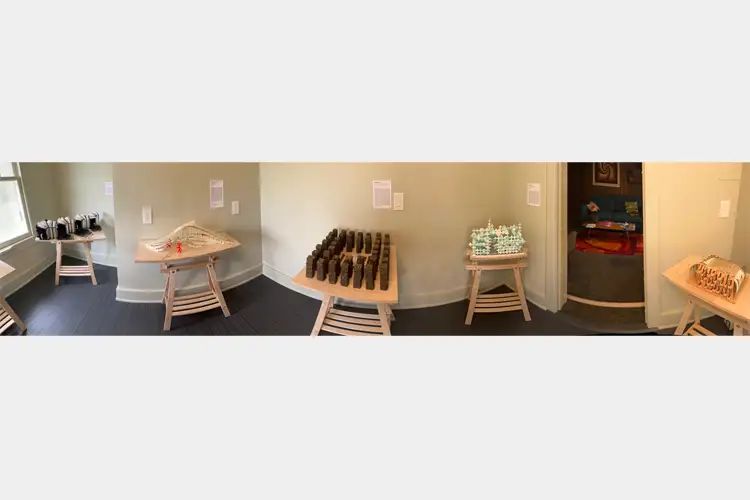 Gallery room with small tabletop sculptures on wooden stands, including dark cylinder pieces, white forms, and geometric models near a doorway. Gallery room with small tabletop sculptures on wooden stands, including dark cylinder pieces, white forms, and geometric models near a doorway.