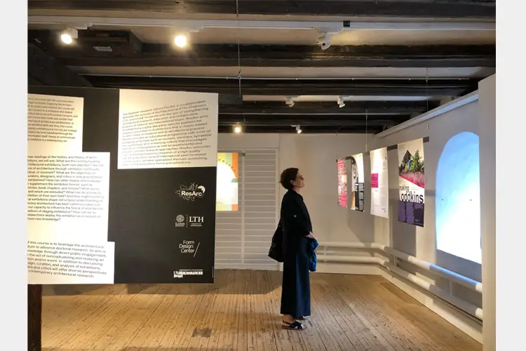 Visitor stands in gallery reading wall text and viewing posters and a large projection in a wooden-floored exhibition space with overhead lighting. Visitor stands in gallery reading wall text and viewing posters and a large projection in a wooden-floored exhibition space with overhead lighting.