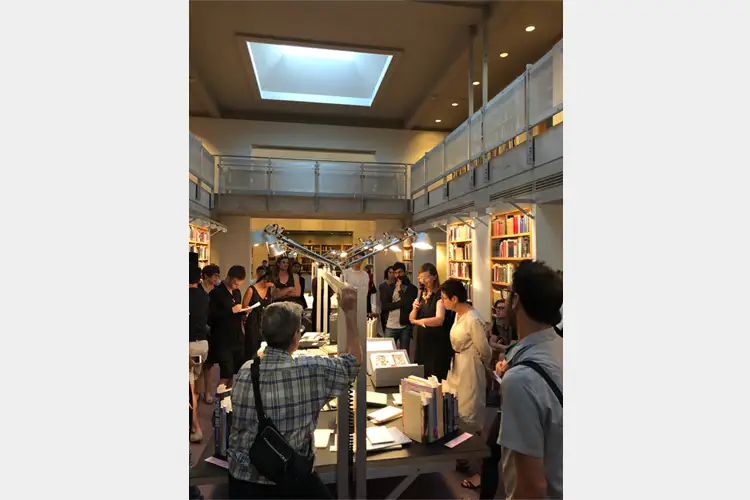 People gather in a library-style gallery around a central table with books and a metal sculpture, viewed from a balcony-lit room with skylight. People gather in a library-style gallery around a central table with books and a metal sculpture, viewed from a balcony-lit room with skylight.