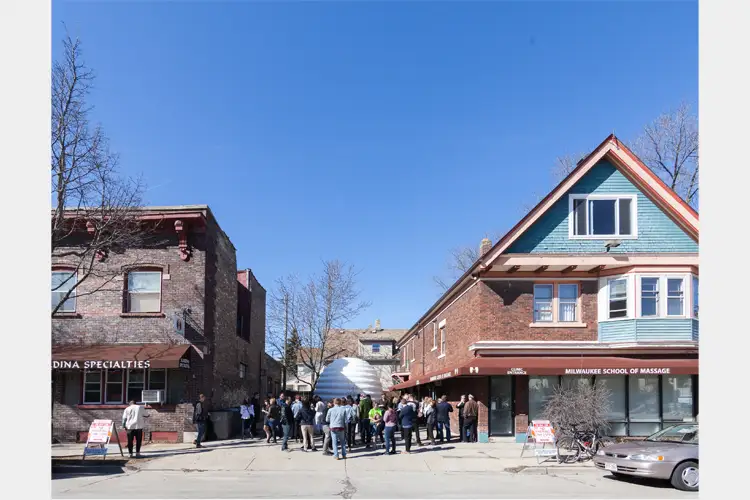 Crowd gathers on a sunny street between brick buildings, with a white dome structure ahead; storefront reads “Milwaukee School of Massage.” Crowd gathers on a sunny street between brick buildings, with a white dome structure ahead; storefront reads “Milwaukee School of Massage.”