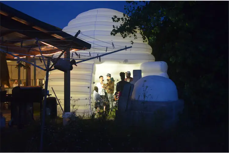 People gathered at night beside a glowing white dome structure, with light illuminating figures standing at the entrance amid trees and outdoor equipment. People gathered at night beside a glowing white dome structure, with light illuminating figures standing at the entrance amid trees and outdoor equipment.