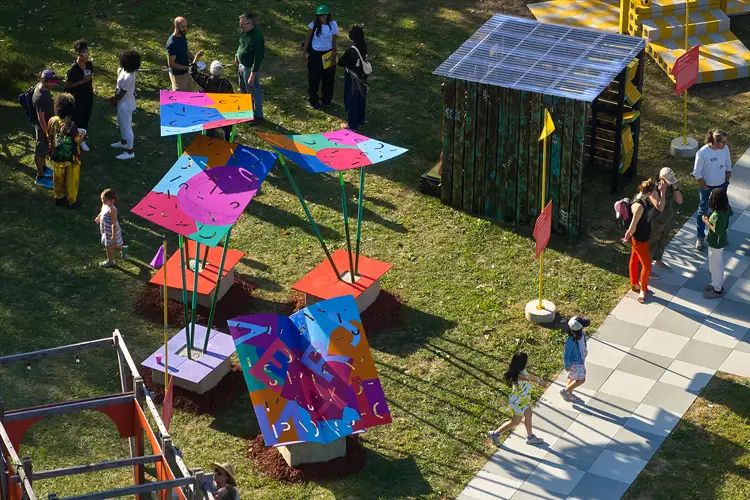 Colorful outdoor installation with painted canopy panels on poles, small shed, and people walking and gathering on grass and pathways. Colorful outdoor installation with painted canopy panels on poles, small shed, and people walking and gathering on grass and pathways.